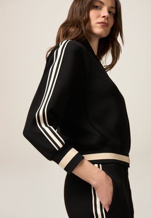Sudadera deportiva negra con mangas largas, presenta tres rayas verticales blancas en cada brazo y un dobladillo acanalado beige.