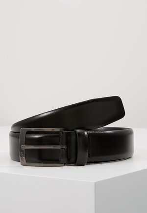 Ceinture en cuir noire avec une boucle en métal argenté, reposant enroulée sur une surface blanche sur un fond clair uni.