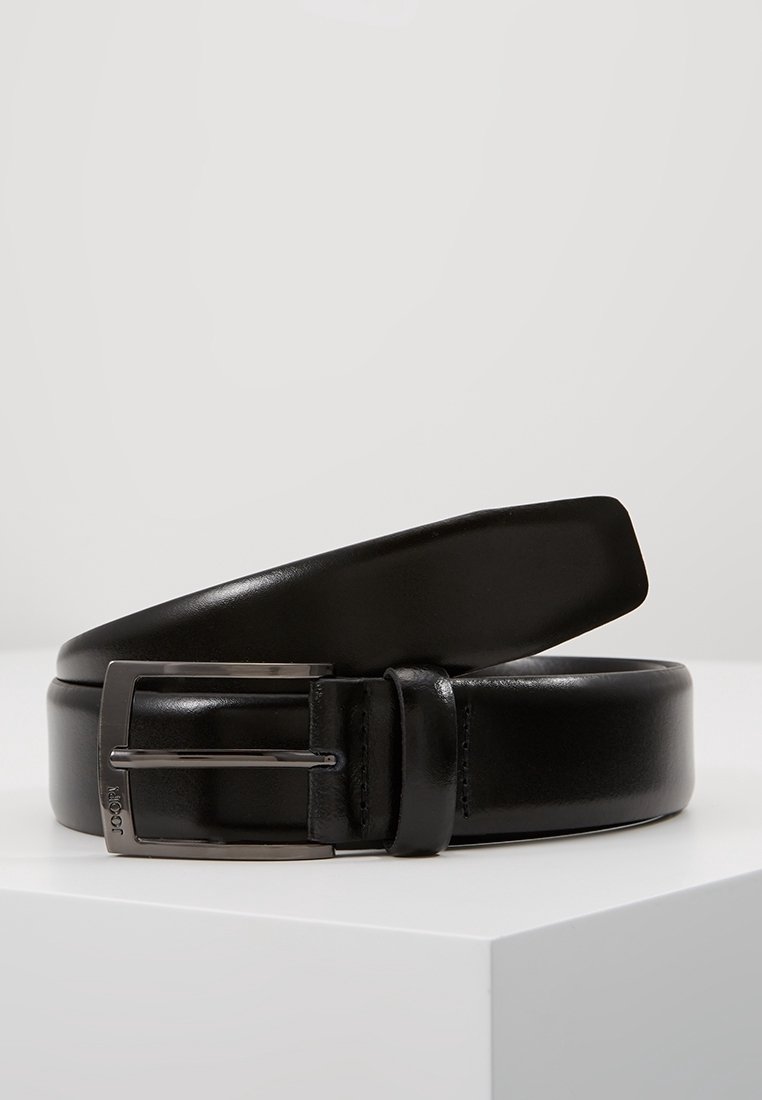 Ceinture en cuir noire avec une boucle en métal argenté, reposant enroulée sur une surface blanche sur un fond clair uni.