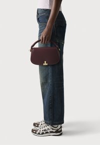Borsa strutturata borgogna con manico superiore, hardware dorato e forma arrotondata, tenuta da una persona che indossa jeans blu e sneaker a fantasia.