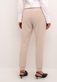Béžové, zúžené kalhoty s hladkým materiálem, elastickými manžetami a dvěma zadními kapsami. V kombinaci s černými střevíci s špičatou špičkou.