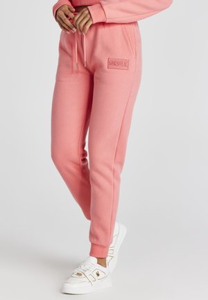 Osoba ubrana w różowe dresowe spodnie SikSilk i białe trampki, stojąca z rękami przy talii na jednolitym tle.