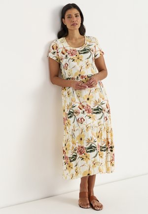 Vrouw staat tegen een witte muur, draagt een witte bloemige short-sleeve midijurk en bruine platte sandalen met twee bandjes.