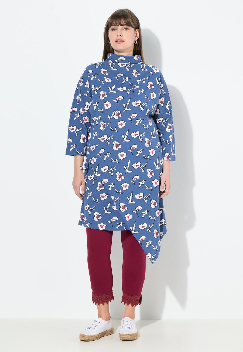Blaue Tunika mit floralem Muster, asymmetrischem Saum und Stehkragen, kombiniert mit burgunderfarbenen Leggings mit Spitzenbesatz am Saum und weißen Sneakers.