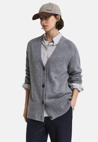 Grauer Strickcardigan mit V-Ausschnitt, Knopfverschluss und geripptem Saum, getragen über einem gestreiften Shirt; kombiniert mit einer hellgrauen Mütze.
