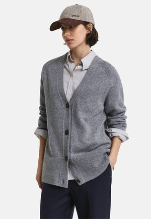 Grijze gebreide cardigan met een V-hals, knoopsluiting en geribde zoom, gedragen over een gestreept shirt; gecombineerd met een lichtgrijze cap.