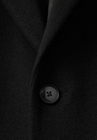 Nærbillede af en sort uldfrakke med spidse revers og en rund sort knap med fire huller, der fremhæver et blødt, tekstureret stof.
