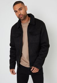 Sort ruskindsjakke med et lammeskindskrage, forbagslommer og lynlås, båret over en beige skjorte i en indendørs setting.