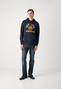 Navy blue hoodie with gold text and emblem, featuring a kangaroo pocket and drawstring hood. Paired with dark denim jeans and black shoes.