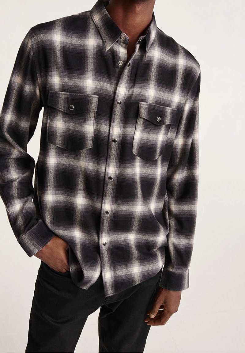 Chemise en flanelle à carreaux noirs et blancs avec col boutonné, deux poches avant et patte de boutonnage, présentée sur un fond sombre.
