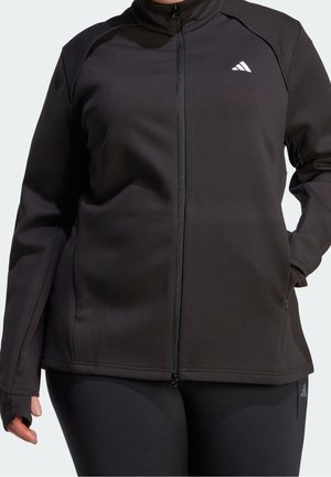 Frau trägt schwarze Sportjacke mit durchgehendem Reißverschluss und Daumenschlaufen, steht mit einer Hand in der Tasche vor einem schlichten hellen Hintergrund.