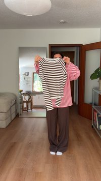 Maglietta a maniche lunghe con righe orizzontali marroni e bianche, colletto a girocollo, tessuto morbido. Tenuta in mano da una persona in una stanza con pavimenti in legno.
