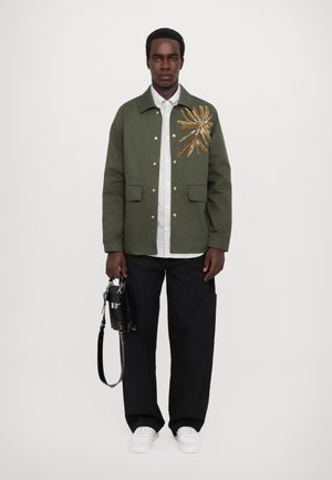 Khaki green jacket with a large gold floral embroidery, featuring snap buttons and front pockets. Paired with black wide-leg pants and white sneakers.