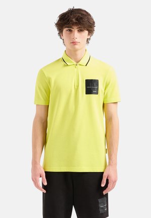 Polo shirt giallo neon con accenti neri, maniche corte e colletto con zip. Presenta una toppa quadrata nera con logo sul petto.