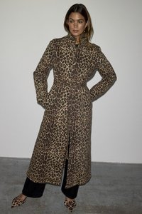 Løveprikket trenchcoat med belte i livet, lange ermer og krage. Bæres over svarte bukser og matchende mønstrede sko.