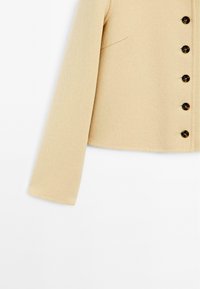 Beige wool blend jacket with a cropped design, featuring a straight sleeve, side pocket, and six dark buttons along the front.