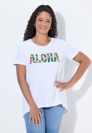 Kobieta z kręconymi włosami, nosząca kolczyki-koła, białą koszulkę z napisem „ALOHA” wykonanym kwiatowym fontem oraz jasnoniebieskie dżinsy, pozująca z jedną ręką na biodrze.