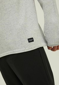 Gros plan d'un pull gris clair texturé avec une petite étiquette noire Jack & Jones près de l'ourlet, porté avec un pantalon noir.