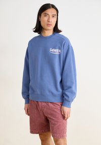 Sweatshirt azul feito de algodão, com um logotipo branco impresso no peito. Combinado com calções de corduroy bordeaux, usados de forma casual.