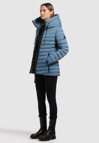 Veste matelassée bleue avec une grande capuche, fermeture éclair et poches latérales. Présente une texture côtelée et une silhouette ajustée. Associée à des leggings noirs et des bottes.