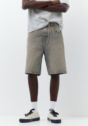 Osoba nosząca szare jeansowe szorty, czarno-białe trampki, białe skarpetki oraz białą i czarną koszulkę w paski ze skrzyżowanymi ramionami.