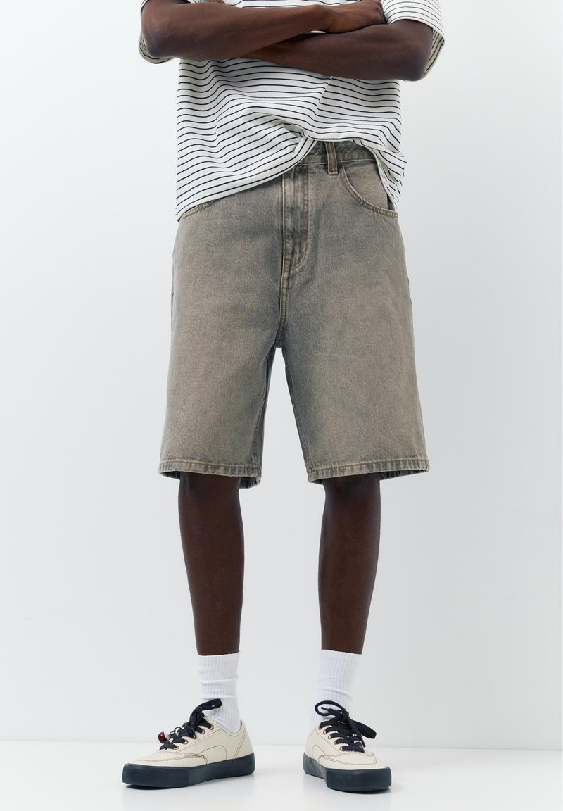 Personne portant un short en jean gris, des baskets noir et blanc, des chaussettes blanches, et une chemise rayée blanche et noire avec les bras croisés.
