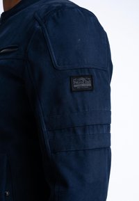 Giacca blu scuro con finitura testurizzata, tasca con zip sul petto e una toppa sul braccio con accenti di cuciture orizzontali.