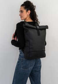 Rucsac negru tip roll-top cu un finisaj texturat, dotat cu bretele ajustabile și o buzunar frontal, purtat de o persoană în ținuta casual.