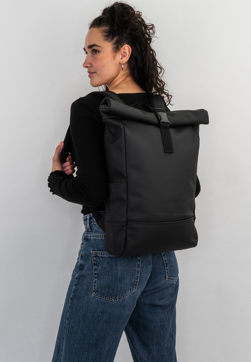 Rucsac negru tip roll-top cu un finisaj texturat, dotat cu bretele ajustabile și o buzunar frontal, purtat de o persoană în ținuta casual.
