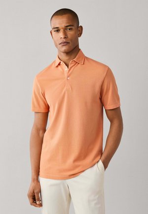 Man draagt een oranje polo met korte mouwen en een witte broek, staand tegen een effen lichtgrijze achtergrond.