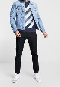 Hellblaue Denimjacke mit Knopfleiste, dunkles navyblaues Grafik-T-Shirt mit blauen und schwarzen diagonalen Streifen, schwarze Hose und weiße Sneaker.