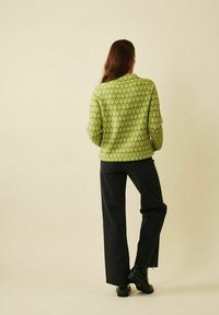 Kvinna med långt brunt hår som bär en grönmönstrad långärmad topp, svarta vida byxor och svarta stövlar, stående mot en enfärgad bakgrund.