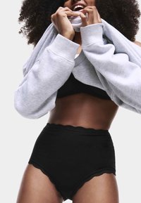 Högmidjade svarta underkläder i mjukt material med spetsdetaljer, kombinerat med en ljusgrå oversized sweatshirt, delvis dragen över.