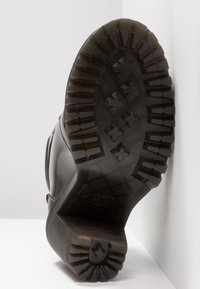 Semelle de botte en caoutchouc noir avec des motifs de crampons profonds et des rainures rectangulaires. Dispose d'un talon prolongé et d'une base texturée pour une meilleure adhérence.