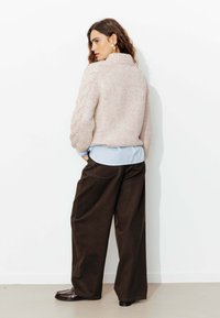 Pull en tricot rose clair avec un col montant et des détails en côtes, associé à un pantalon large marron foncé et des chaussures noires.