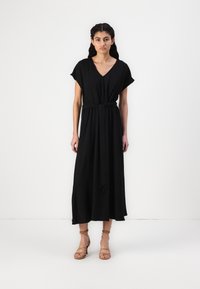 Black maxi dress with short sleeves, v-neck, cinched waist, and flowing skirt; smooth fabric drapes softly; paired with strappy beige sandals.