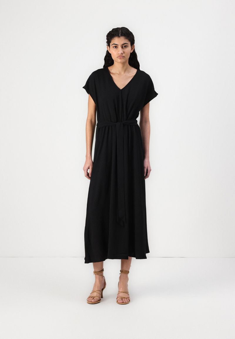 Black maxi dress with short sleeves, v-neck, cinched waist, and flowing skirt; smooth fabric drapes softly; paired with strappy beige sandals.