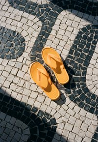 Paire de tongs orange placée sur un pavement en mosaïque avec des motifs géométriques noirs et blancs à la lumière naturelle du soleil.
