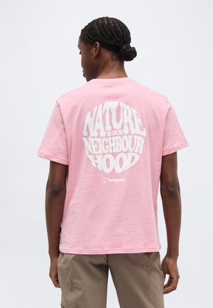 Osoba s opletenými vlasmi nosí ružové tričko s bielym nápisom "Príroda je naše susedstvo" a béžové nohavice, stojaca chrbtom k jednoliatemu pozadiu.