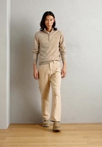 Pulover bej tricotat cu fermoar pe un sfert, asortat cu pantaloni bej deschis și încălțăminte bej asortată, pe un fundal gri neutru.