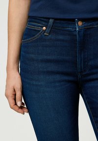 Jean en denim bleu foncé avec un léger effet de décoloration et des coutures orange, dotés d'un bouton en bronze et de détails de poches avant.