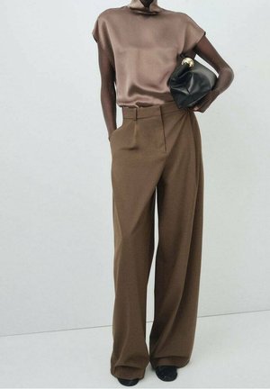 Mannequin portant un haut sans manches à col roulé marron et un pantalon marron à jambes larges, tenant une pochette noire sur un fond clair uni.