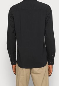 Camicia nera a maniche lunghe con colletto a bottoni, caratterizzata da una texture liscia e un orlo curvo. Bottoni ai polsi e dettaglio sul retro.