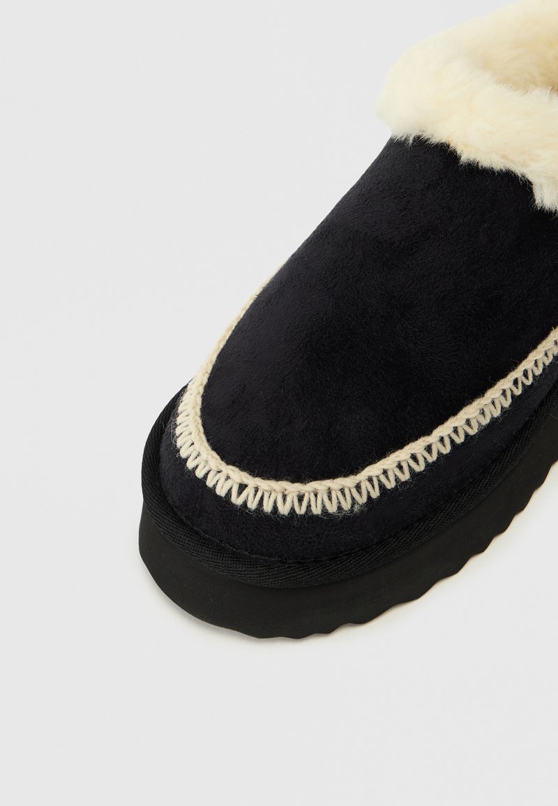 Pantofole in suede nero con fodera in pelliccia sintetica, caratterizzate da un bordo cucito color crema e una suola in gomma nera testurizzata.
