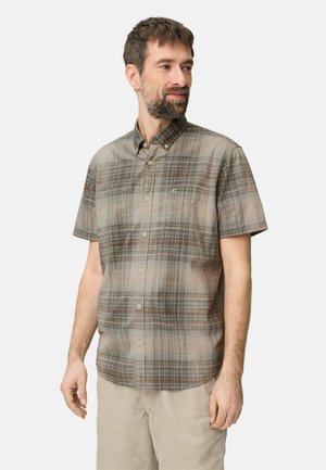 Mężczyzna z krótkimi brązowymi włosami i brodą, ubrany w brązowo-szarą kraciastą koszulę z krótkim rękawem na guziki oraz jasnobeżowe spodnie, patrzy w prawo.