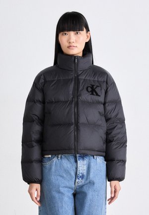 Cazadora tipo puffer negra con cuello alto y cierre frontal, con una superficie texturizada y el logo bordado de "CK" en el lado izquierdo del pecho.