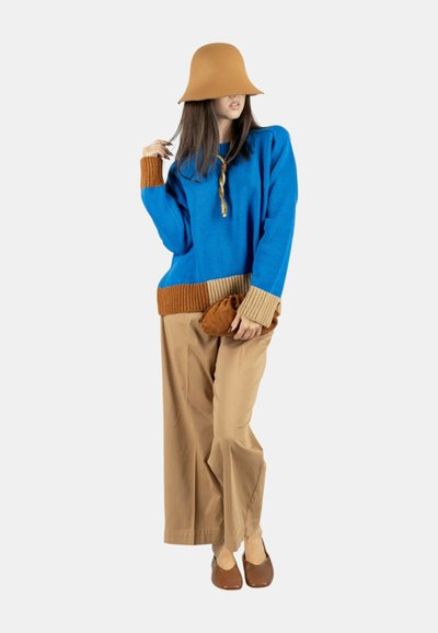 Maglione a maglia blu con polsini a costine in contrasto marrone e beige, pantaloni ampi color cuoio, scarpe marroni e cappello coordinato. Pochette marrone in mano.