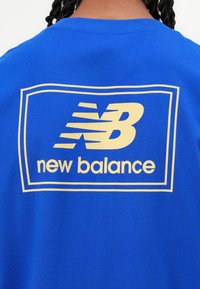 Blå bomulds t-shirt med et stort gult "NB" logo og "new balance" tekst inden i en rektangulær ramme på ryggen.