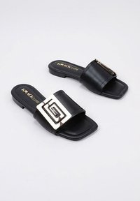 Sandalias negras con una correa ancha que presenta una hebilla geométrica plateada. Suela plana y textura suave. Nombre de la marca "KRAKCORE" impreso en dorado.