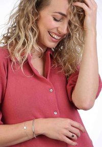 Button-up shirt in pink fabric, featuring a collar and short sleeves. Silver bracelets adorn a wrist, with a minimalist design and heart charm.